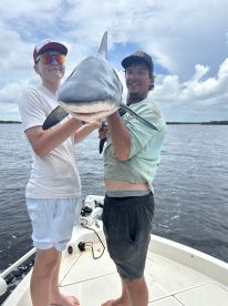 Two anglers proudly displaying a large shark while fishing from a boat in a coastal waterway.