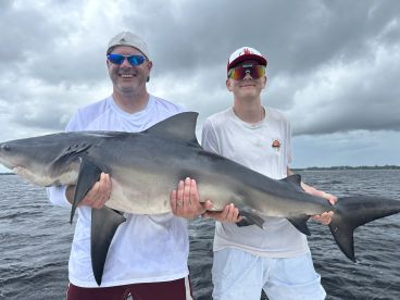 A proud angler and a young fisherman displaying a large shark caught during their fishing trip, set against a cloudy backdrop on the water.