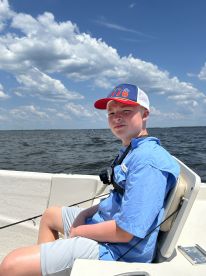 A young angler seated in the boat, enjoying a sunny day on the water, with a clear view of the sky and lake.