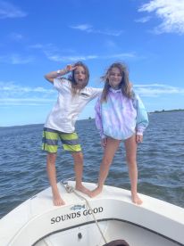 Two young girls posing playfully on the bow of a boat named 'Sounds Good' while enjoying a sunny day on the water.