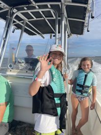 Two young girls wearing life jackets and smiling while enjoying a boat ride, with a captain visible in the background.