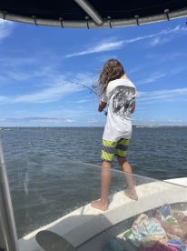 A young angler actively reeling in a fish while standing on the bow of a boat, enjoying a sunny day on the water.