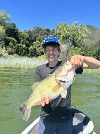 Young angler proudly holding a large Largemouth Bass while fishing on a serene lake surrounded by lush greenery.