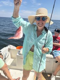 A woman proudly displaying a Red Snapper while fishing on a boat, with a child visible in the background, enjoying a sunny day on the water.