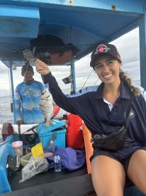 A young woman proudly displaying her catch, a small fish, while fishing on a colorful boat in open waters.