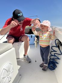A young child joyfully holds a small fish while an adult assists on a fishing boat, showcasing a fun family fishing experience.