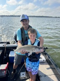 A young boy proudly displays a Walleye he caught while fishing with an adult on a boat, showcasing a fun day on the water.