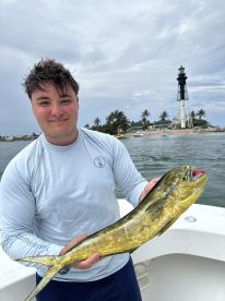 A young angler proudly displaying a vibrant Mahi Mahi while fishing near a lighthouse, showcasing a successful day on the water.