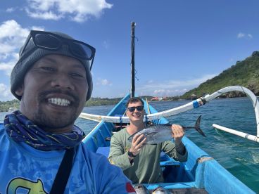 Two anglers on a traditional fishing boat proudly displaying their catch, a large fish, while enjoying a sunny day on the water.