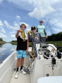 Three young anglers proudly displaying their catches of Largemouth Bass while fishing from a boat in a calm waterway.