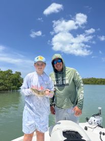 A young angler proudly displaying a caught flounder while fishing with an adult companion on a sunny day in a coastal environment.