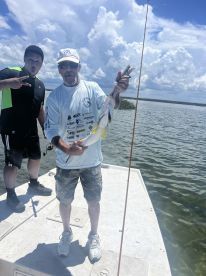 Two anglers on a boat, one proudly displaying a caught fish while the other poses playfully, showcasing a fun day of fishing in a scenic waterway.