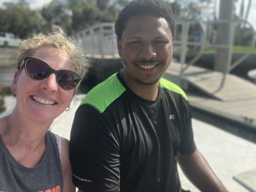 Two individuals smiling for a selfie on a boat, with a dock visible in the background.