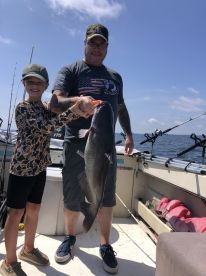 A young angler and an adult proudly displaying a large catfish while fishing on a boat, enjoying a sunny day on the water.
