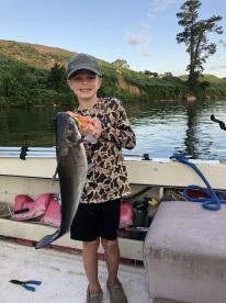 A young angler proudly displaying a large catfish while fishing on a calm river, surrounded by lush greenery.
