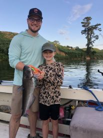 A young boy and an adult proudly displaying a large Catfish while fishing on a river, showcasing a memorable family fishing experience.