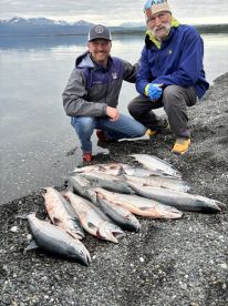 Two anglers proudly displaying a catch of multiple Salmon on a rocky shore, with a scenic backdrop of mountains and water.
