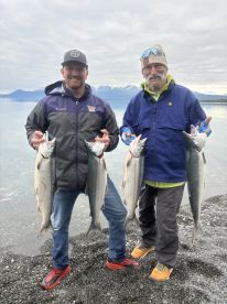 Two anglers proudly displaying their catch of Silver Salmon on a rocky beach with a scenic mountain backdrop.