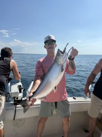 Angler proudly displaying a large Salmon while fishing on a sunny day, with fellow fishermen in the background.