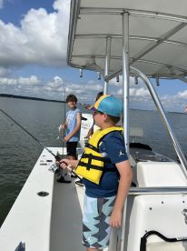 Two young anglers fishing from a boat, enjoying a sunny day on the water, with one holding a fishing rod and the other smiling in the background.