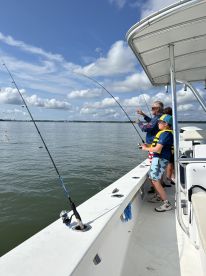 A family enjoying a fishing trip on a boat, with a child in a life jacket actively reeling in a catch while the adult assists. The calm waters and clear skies create a perfect fishing environment.