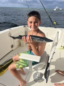 A young angler proudly displaying a Barracuda while seated on a fishing boat, with a scenic ocean backdrop.