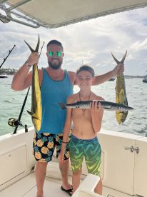 A proud father and son duo displaying their catch of Mahi Mahi and Barracuda while enjoying a day of fishing on a boat.