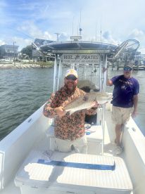 Angler proudly displaying a large Redfish aboard a charter boat, with a scenic waterfront in the background.