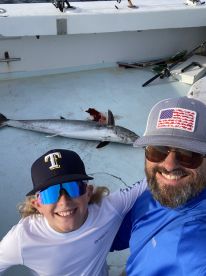 A young angler and an adult proudly pose for a selfie with a large fish on the deck of a fishing boat, showcasing their successful catch.