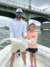A young girl and an adult proudly display a caught shark while on a boat, showcasing a successful fishing trip.