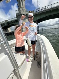 A mother and daughter duo proudly displaying their catch, a small fish, while fishing on a boat near a bridge.