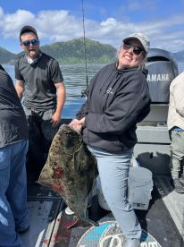 Angler proudly displaying a large Halibut while fishing in a scenic Alaskan environment, with mountains in the background.