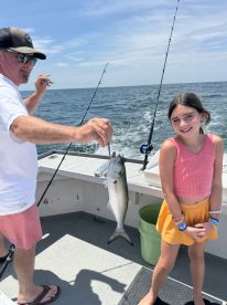 A young girl and an adult are on a boat, with the adult proudly displaying a caught fish, likely a Bluefish, during a sunny day on the water.