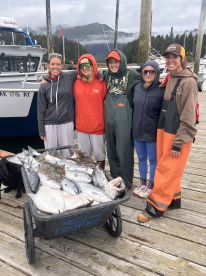 A group of five anglers proudly displaying their catch of various fish in a wheelbarrow at the dock, showcasing a successful fishing trip in a scenic coastal environment.