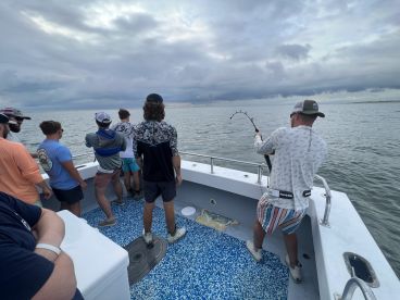 Group of anglers actively fishing on a charter boat, with one person reeling in a catch against a cloudy backdrop.