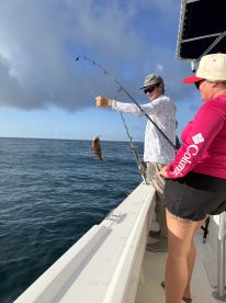 A young angler is reeling in a fish while fishing off the coast, with a female companion observing the action.