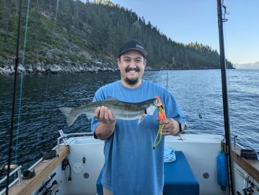 Angler proudly displaying a caught trout while fishing from a boat in a scenic lake surrounded by forested hills.