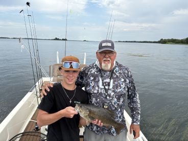 A young angler proudly displaying a large Largemouth Bass while fishing on a boat, accompanied by an experienced guide in a scenic lake setting.