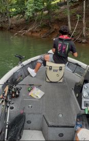 A young angler seated on a YETI cooler, preparing to fish on a serene lake, surrounded by fishing gear and a lush forest backdrop.
