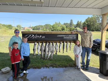 A family poses proudly in front of a catch board displaying several fish after a successful fishing trip in Manistee, MI.