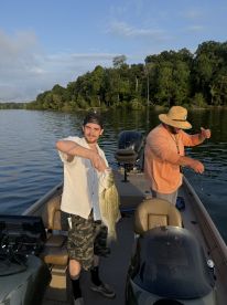 A young angler proudly displaying a large striped bass while fishing on a serene lake, with lush greenery in the background.