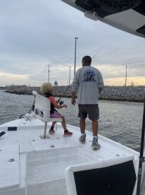 A young angler in a life jacket is seated on a boat, preparing to fish under the guidance of an adult, with a scenic backdrop of the water and sky.