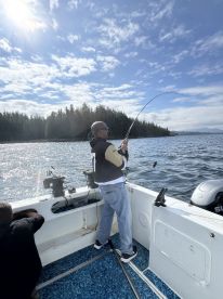 An angler actively reeling in a fish while fishing on a boat surrounded by scenic forested coastline.