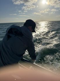A lone angler is reeling in a catch while fishing on a boat, with the sun setting over the water.