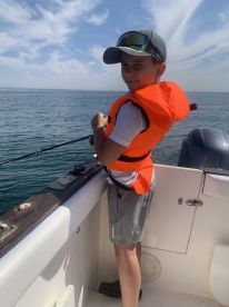 A young angler in an orange life jacket is casting a line from the side of a boat on a sunny day, showcasing the excitement of fishing in open waters.