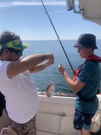 Two young anglers engaged in fishing on a boat, with one holding a freshly caught Snapper while the other assists in reeling in.