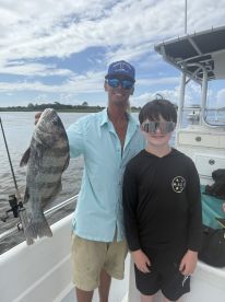 A proud young angler and an adult holding a large Black Drum on a fishing charter, showcasing their catch against a scenic water backdrop.