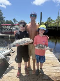 Two young anglers proudly displaying their catches, a large Black Drum and a smaller fish, on the dock after a successful fishing trip.