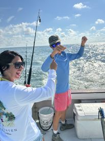 A young angler and a companion are actively fishing on a boat, with the sun shining and a beautiful ocean backdrop.