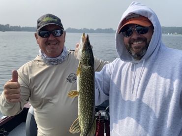 Two anglers proudly displaying a Northern Pike while fishing on a calm lake, showcasing a successful day on the water.
